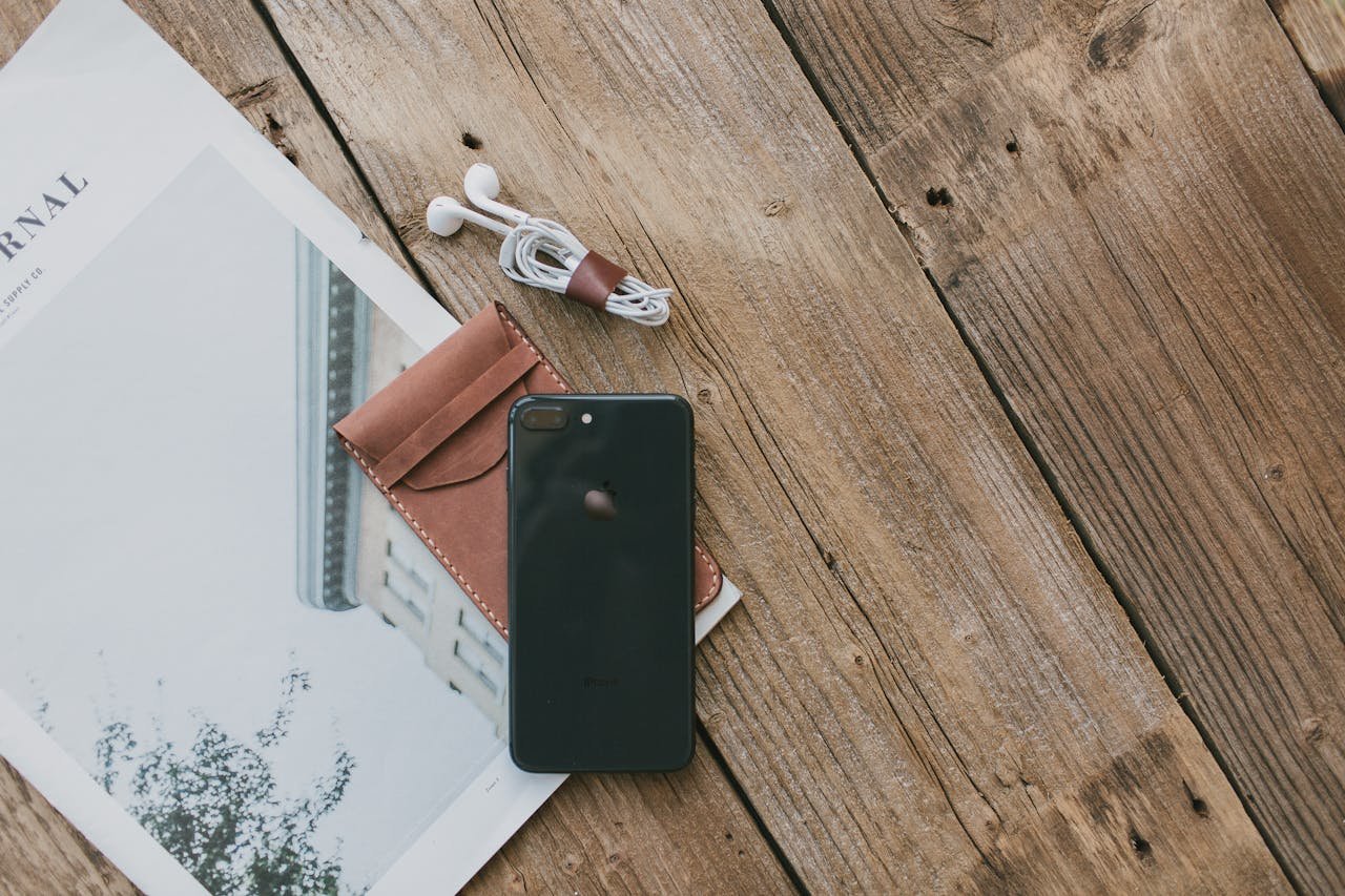 Flat lay photo of smartphone, earphones, leather case, and journal on a wooden surface.